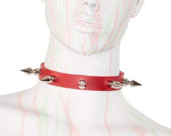 May include: A red leather choker necklace with silver-colored spikes and studs. The choker is displayed on a white mannequin head with red and green paint streaks.