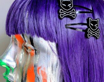 May include: Two black hair clips featuring a white cat skull and crossbones design. Each clip has a stylized feline head with pointed ears and a small star detail above the eye, positioned on vibrant purple hair with a silver and orange mannequin head.