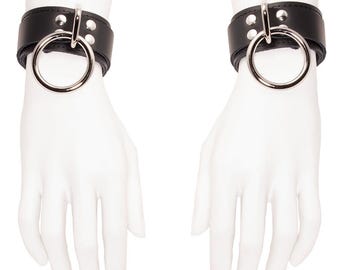 May include: A pair of black leather wrist cuffs with silver-colored metal rings. Each cuff has a wide band with three silver rivets. The cuffs are displayed on white mannequin hands against a white background.