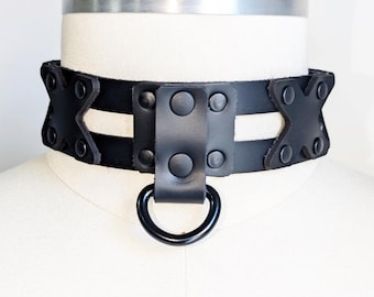 May include: A black leather collar with a D-ring and rivet details. The collar features a geometric design with cut-out sections and is displayed on a mannequin head.