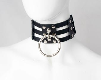 May include: A black leather choker with three horizontal straps and a large silver ring. The choker is secured with silver rivets and is displayed on a white mannequin head. The design is bold and edgy.