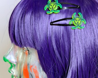May include: Two black metal hair clips featuring bright neon green biohazard symbol charms. The charms have a translucent, layered appearance with dark green outlines, securely attached to the top of each standard snap-style hair clip.