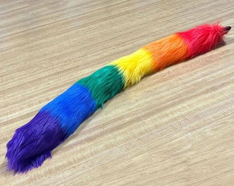 May include: A long, fluffy tail in a rainbow of colors, including purple, blue, green, yellow, orange, and red. The tail is made of faux fur and is lying on a light-colored wooden surface. The tail is a fun accessory.