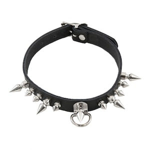 Könnte beinhalten: Schwarze Leder-Halskette mit silberfarbenen Kegel-Nieten und einem Metallring. Die Halskette hat einen Schnallenverschluss und ist für das Tragen um den Hals konzipiert. Der Stil ist kantig, möglicherweise gothisch oder punkig.