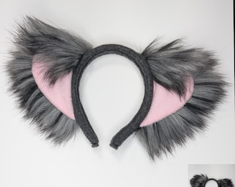 May include: A headband featuring two large, pointed animal ears. The ears are covered in long, shaggy gray faux fur with black tips, while the inner portions are lined with smooth, light pink fabric. The headband itself is wrapped in dark gray fabric.