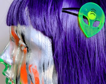 May include: A neon green acrylic hair clip shaped like an alien head with a black metal slide bar. The alien design features a circular forehead detail and two almond-shaped eyes. The hair clip is attached to a head of vibrant purple hair.