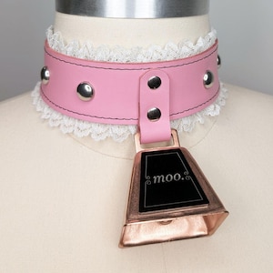 Può includere: Choker in pelle rosa con borchie argentate e rifiniture in pizzo bianco. È attaccata una campana color rame con la scritta "moo.". Il choker è esposto su un manichino.