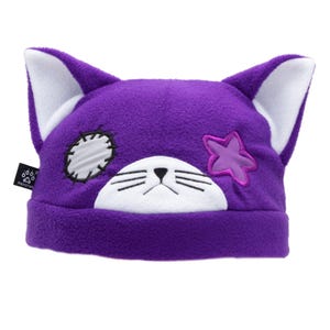 May include: Purple cat-eared beanie hat with white inner ears. The hat features a white face with embroidered whiskers and a black nose. A pink star and a white patch with black stitching are on the hat. The brand name "PAWSTAR" is on a black tag.
