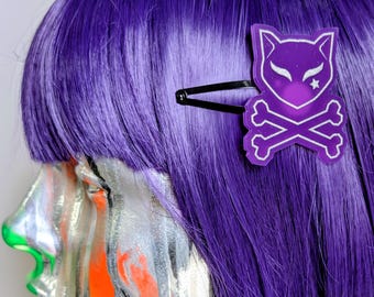 May include: A black metal hair clip featuring a purple acrylic charm shaped like a cat head above crossed bones. The charm has a glittery finish with white outlines, a small white star detail, and pointed ears, attached to a standard snap hair clip.