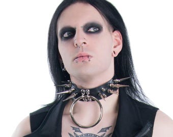 May include: Black leather choker with silver spikes and a large silver ring. The choker is a bold accessory, perfect for alternative fashion. The design features multiple silver spikes and a prominent ring in the center.