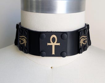 May include: Black leather choker with gold ankh and eye of Horus symbols. The choker is on a mannequin and features a rectangular center piece with the ankh symbol and smaller side pieces with the eye of Horus symbol.