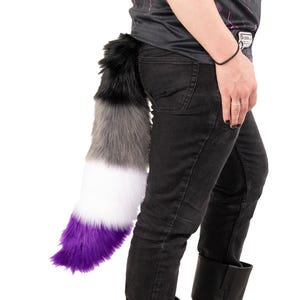 May include: A fluffy, multi-colored tail accessory. The tail features a gradient of colors, starting with black at the top, transitioning to gray, white, and ending with a vibrant purple. The tail is attached to a person wearing black jeans.