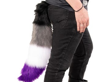 May include: A fluffy, multi-colored tail accessory. The tail features a gradient of colors, starting with black at the top, transitioning to gray, white, and ending with a vibrant purple. The tail is attached to a person wearing black jeans.