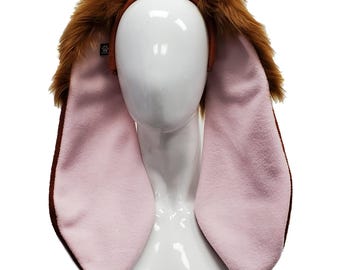 May include: A bunny ear headband with long, pink inner ears and brown outer edges. The headband features fluffy brown fur around the ears and a brown band. The ears are displayed on a white mannequin head.