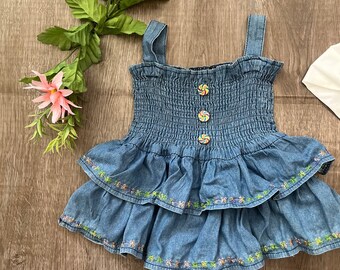 May include: A blue denim romper with a tiered skirt and spaghetti straps. The bodice is smocked, and the skirt is adorned with colorful floral embroidery. The romper features three candy-colored buttons and is suitable for a baby or toddler.