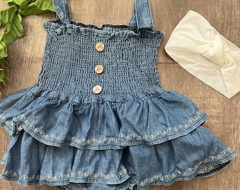 May include: A denim baby romper with a ruffled skirt and embroidered detailing. The romper features a smocked bodice, three decorative buttons, and snap closures. A white knotted headband is included. A pink flower and greenery are in the background.