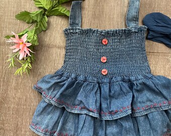 May include: A blue denim baby romper with a smocked bodice and tiered skirt, featuring three pink buttons and pink stitching. Includes a matching blue headband. The outfit is designed for a baby or toddler.