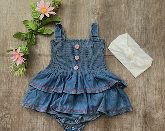 May include: Blue denim baby romper with a tiered skirt and red stitching. The romper has a smocked bodice, flower-shaped buttons, and snap closures. A white knotted headband is included. The outfit is displayed on a wooden surface.