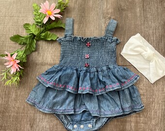 May include: A denim baby romper with a tiered skirt and pink embroidered details. The romper has a smocked bodice and button closures. A white knotted headband and pink flowers are also in the image.