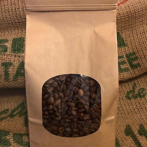 May include: A brown paper bag with a clear oval window displaying roasted coffee beans. The bag has a zip closure and is set against a burlap sack background.