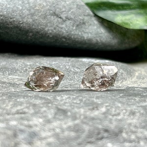 May include: A pair of clear quartz crystal earrings with a faceted, geometric shape. The earrings are set against a gray background.
