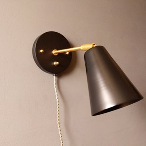 Op de afbeelding: Zwarte wandlamp met een gouden arm en een zwarte, kegelvormige kap. De lamp heeft een snoer met een gevlochten stoffen bekleding.