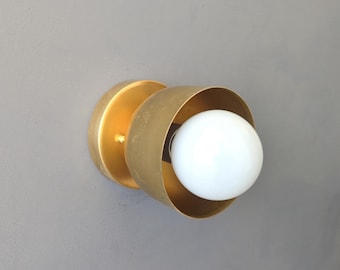 May include: A modern wall sconce with a brass finish. The fixture features a circular backplate and a curved shade that partially surrounds a white globe light bulb. The sconce is designed for indoor use and adds a touch of contemporary style.