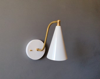 May include: A white cone-shaped wall sconce with a brass arm and accents. The sconce is mounted on a white circular base. The fixture is against a gray wall, showcasing a modern design.