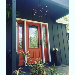 May include: A red front door with a white trim and a glass panel. The door has a black door handle and a black door knocker. The door is surrounded by a dark gray wall. The house number 2465 is visible on the wall. A black starburst chandelier hangs above the door.