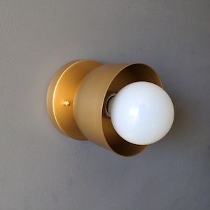 A modern and minimalist style wall sconce with half spherical shaped shade, flat wall base, bright metallic gold finish, white globe bulb. Gray background.