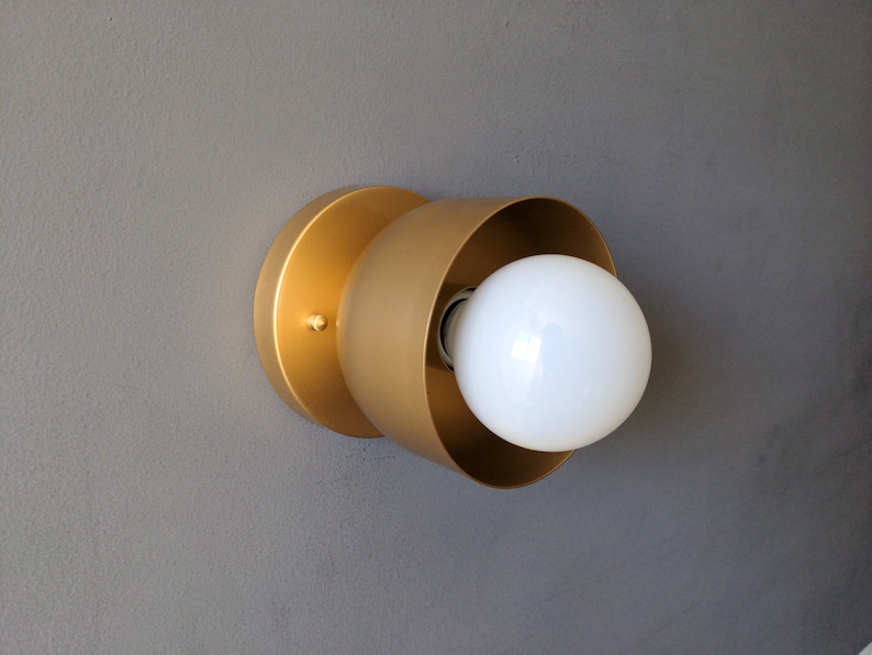 A modern and minimalist style wall sconce with half spherical shaped shade, flat wall base, bright metallic gold finish, white globe bulb. Gray background.