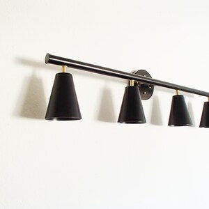 May include: A black metal light fixture with four black cone-shaped shades. The fixture is mounted to a white wall.