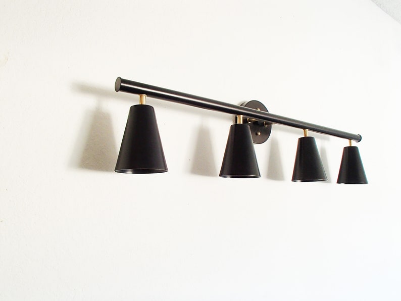May include: A black metal light fixture with four black cone-shaped shades. The fixture is mounted to a white wall.