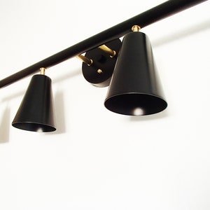 May include: A black metal light fixture with four black cone-shaped shades. The fixture has a gold finish and is mounted to a white wall.