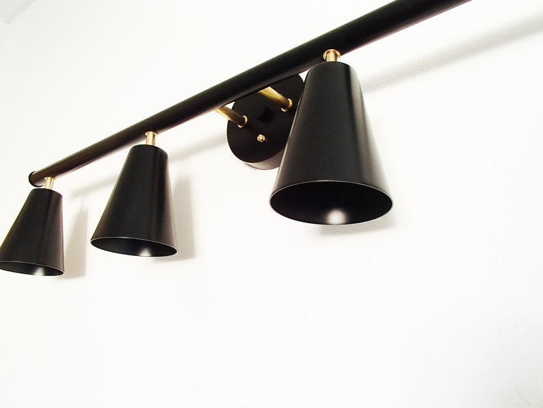 May include: A black metal light fixture with four black cone-shaped shades. The fixture has a gold finish and is mounted to a white wall.