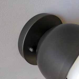 A modern and minimalist style wall sconce with half spherical shaped shade, flat wall base, black finish, white globe bulb. Gray background. Up close detailed photo.