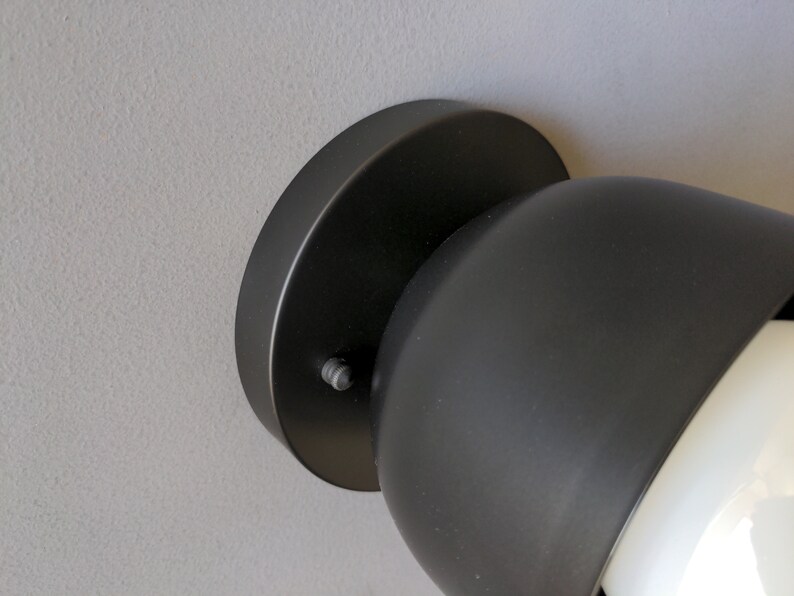 A modern and minimalist style wall sconce with half spherical shaped shade, flat wall base, black finish, white globe bulb. Gray background. Up close detailed photo.
