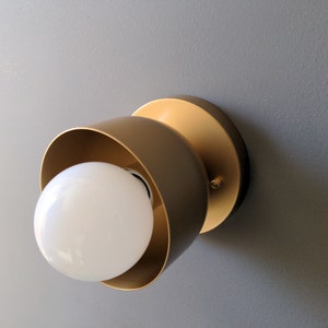 A modern and minimalist style wall sconce with half spherical shaped shade, flat wall base, bright metallic gold finish, white globe bulb. Gray background.