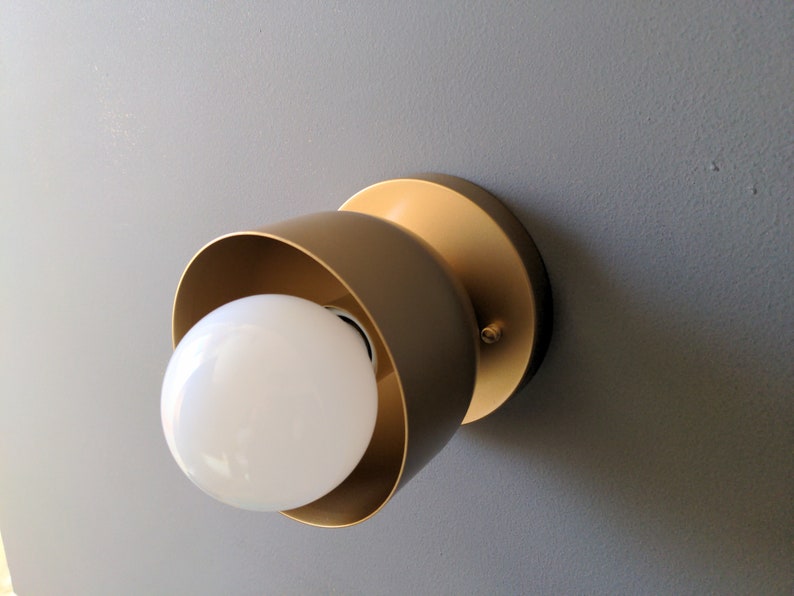 A modern and minimalist style wall sconce with half spherical shaped shade, flat wall base, bright metallic gold finish, white globe bulb. Gray background.