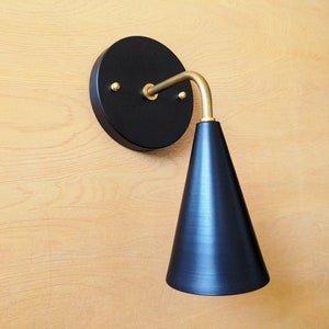 Lighting sconce, black wall base and narrow tapered cone shade, with gold brass 90 degree curved stem and mounting hardware. Wood grain background.