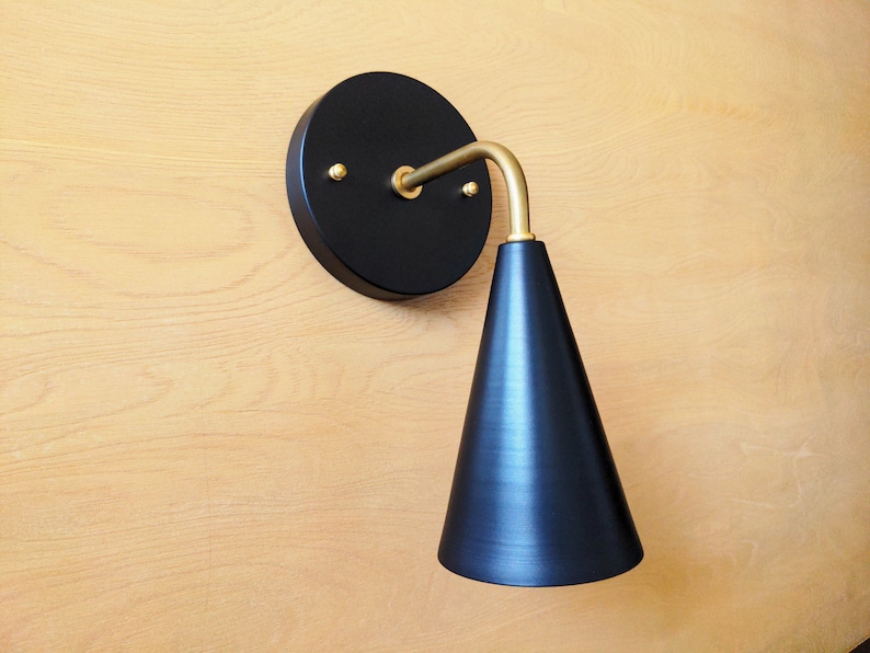 Lighting sconce, black wall base and narrow tapered cone shade, with gold brass 90 degree curved stem and mounting hardware. Wood grain background.