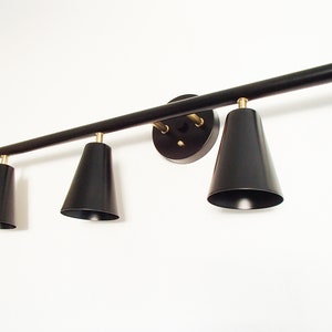 May include: Black track lighting fixture with three black cone-shaped shades. The fixture is mounted to a white wall.