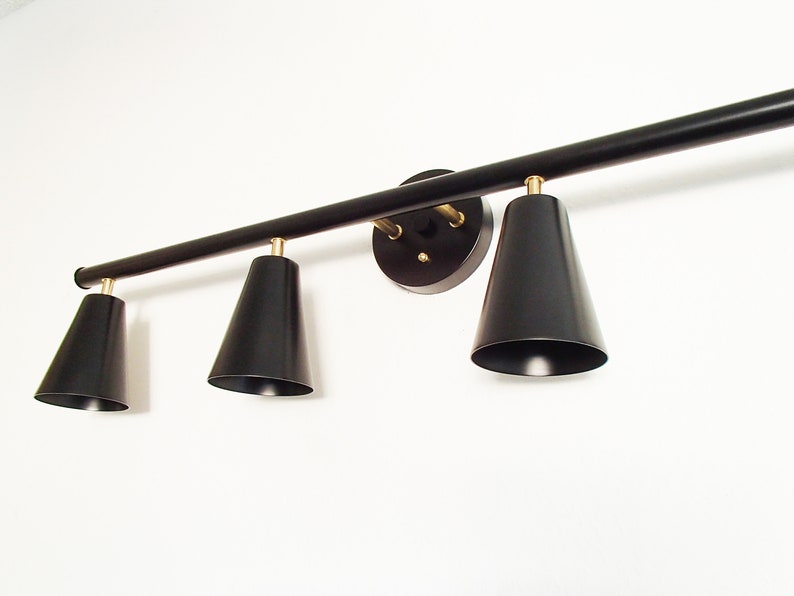 May include: Black track lighting fixture with three black cone-shaped shades. The fixture is mounted to a white wall.