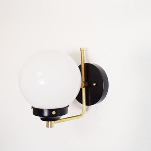 Peut inclure: Une applique murale noire et en laiton avec un globe lumineux blanc. L'applique a une base ronde noire et un bras en laiton qui s'&eacute;tend jusqu'au globe.