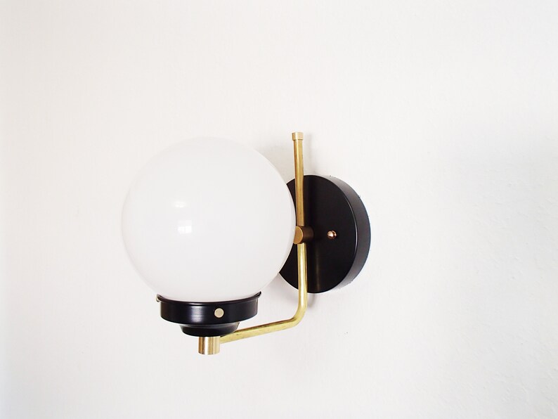 Peut inclure: Une applique murale noire et en laiton avec un globe lumineux blanc. L'applique a une base ronde noire et un bras en laiton qui s'&eacute;tend jusqu'au globe.