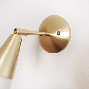 Satin brass finished wall lighting sconce. Round wall base, stem with swivel and small conical shade angled downwards. White globe bulb fitted. White background. Close up view.