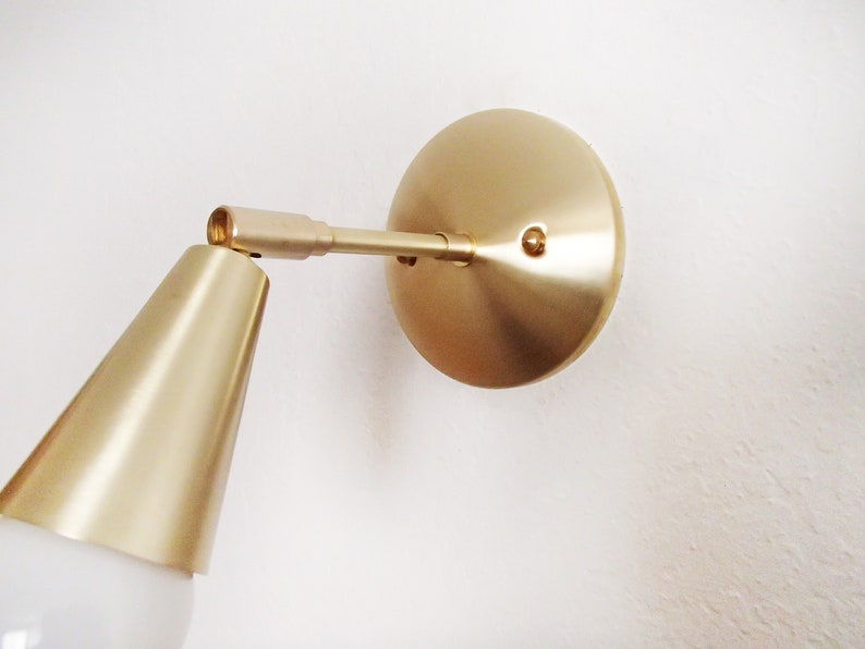 Satin brass finished wall lighting sconce. Round wall base, stem with swivel and small conical shade angled downwards. White globe bulb fitted. White background. Close up view.