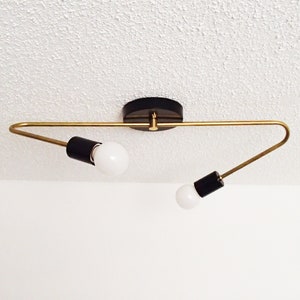 May include: A modern ceiling light fixture with a black base and two arms in a gold finish. Each arm holds a black cylindrical socket with a white bulb. The fixture is mounted on a white ceiling.
