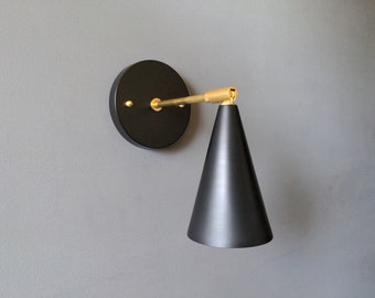 May include: A black wall sconce with a gold arm and a black cone-shaped shade. The sconce is mounted on a circular base.