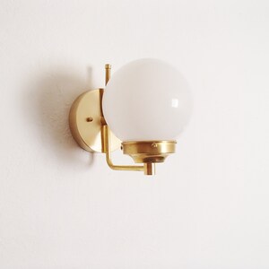 Peut inclure: Une applique murale dor&eacute;e avec un globe lumineux blanc. L'applique a une base ronde et un bras incurv&eacute;.